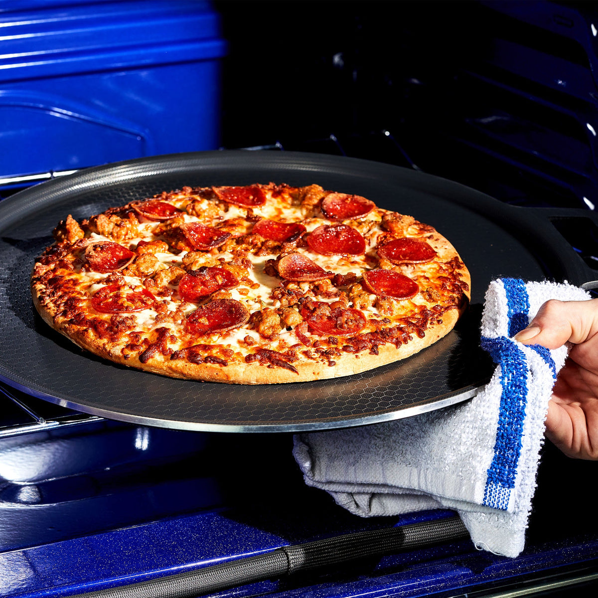 A hand taking a pepperoni pizza out of the oven on the HexClad hybrid pizza steel